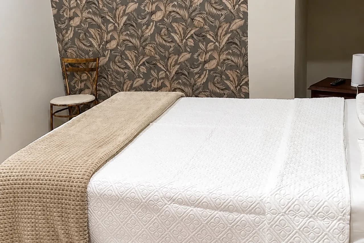 Cama de casal da Suíte Cascata, com colcha branca e manta bege, diante de parede com papel de parede floral