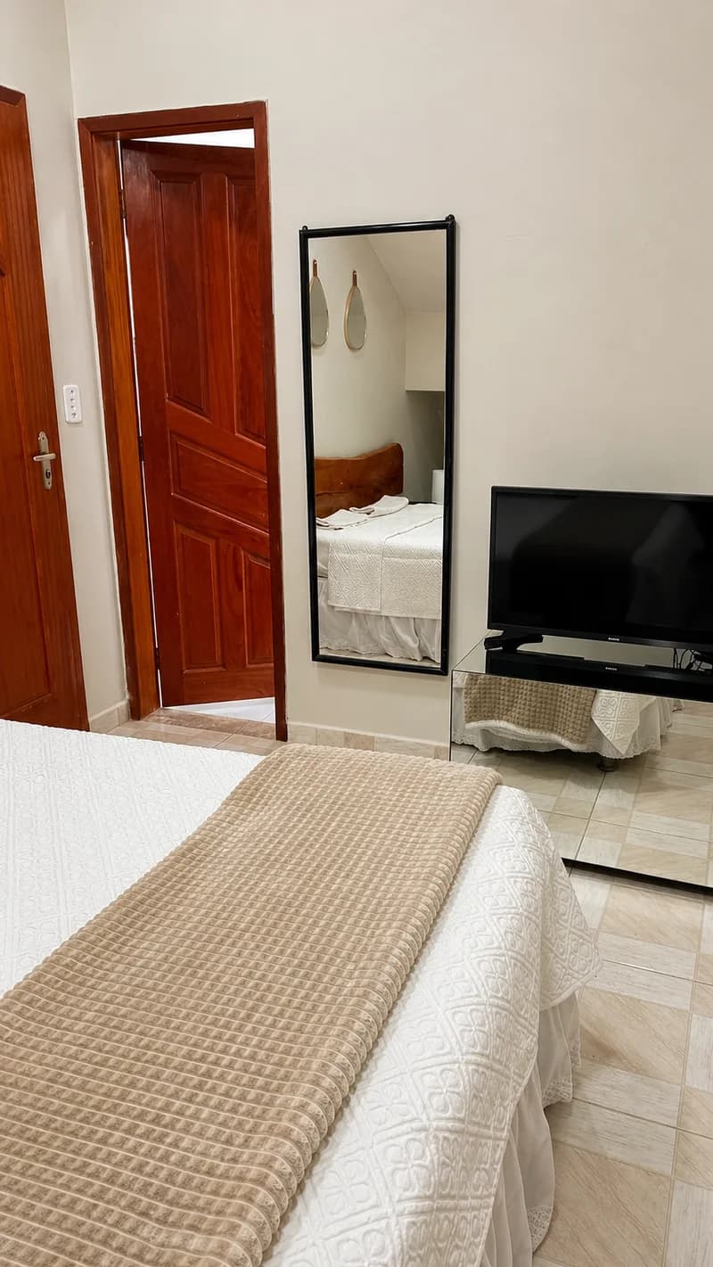 Vista do quarto da Suíte Cascata, com espelho de corpo inteiro, TV e porta de madeira