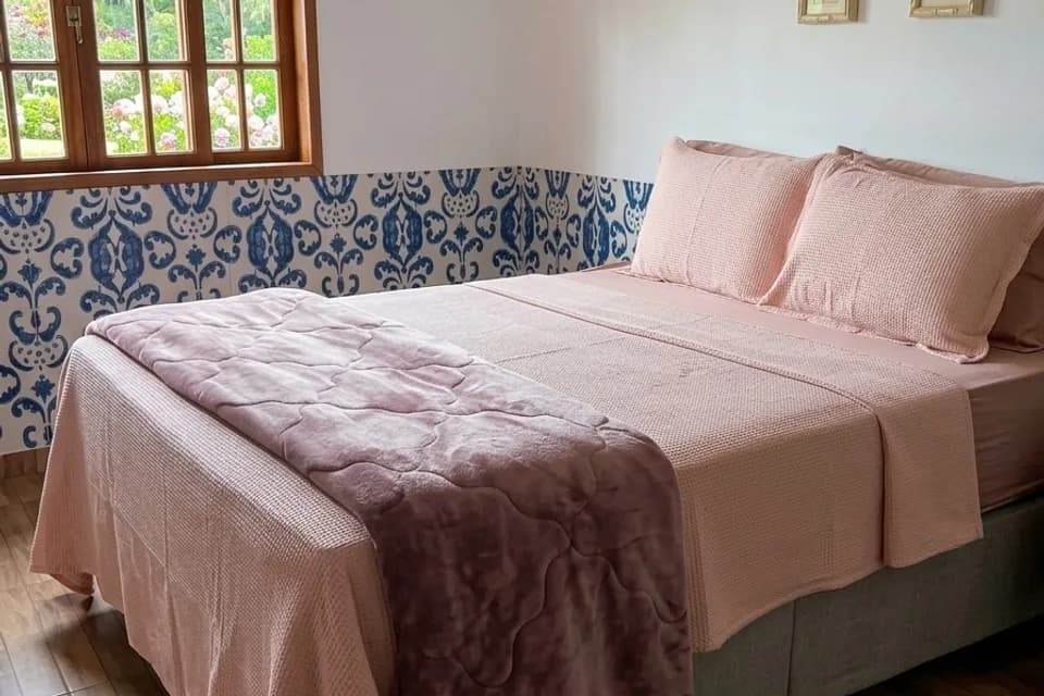 Cama de casal da Suíte Ipê, com roupa de cama rosada e faixa de azulejos azuis na parede
